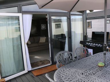 An outdoor patio with a metal table and chairs under an umbrella next to a gas grill and open glass doors leading to a living room with a sofa at Tiggywinkles in Chipping Norton