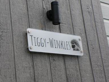 A white sign with the text tiggy winkles and an image of a hedgehog on a gray wooden wall