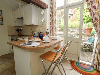A kitchen with a wooden breakfast bar set with cups and pastries and two stools next to a glass door leading to a garden at The Elbow in Frampton