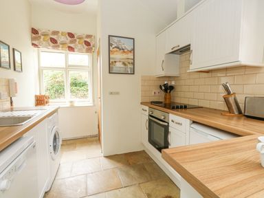 A kitchen with white cabinets wooden countertops a washing machine oven stovetop window with patterned curtain and tiled floor at The Elbow in Frampton