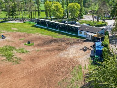 An outdoor driving range with covered golf practice bays and parked cars near trees at The Red in Llanerch Park near St Asaph