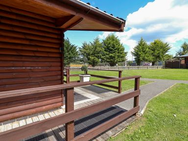 A view of a log cabin with a wooden railing and grass area at The Roe in Llanerch Park near St Asaph