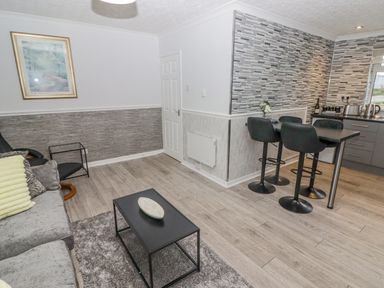 A living area with a gray sofa a black coffee table a small side table and a kitchen area with black stools and a tiled wall at 48 Gower Holiday Village in Scurlage