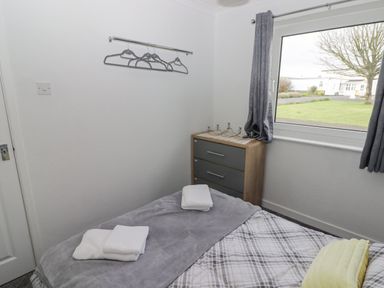 A bedroom with a bed covered by a gray blanket and folded towels a chest of drawers by a window with gray curtains and coat hangers on the wall at 48 Gower Holiday Village in Scurlage