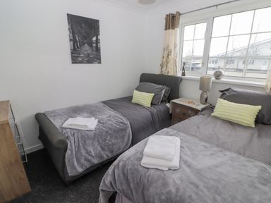 A bedroom with two single beds with grey bedding and yellow pillows a bedside table with a lamp and window with curtains at 48 Gower Holiday Village in Scurlage