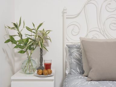 A bedside table with a glass vase of green leaves a lamp a glass of red juice and a plate with three pears next to a bed with pillows at Bushwood Lodge in Wendron near Helston
