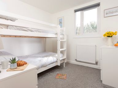 A bedroom with white bunk beds a window a radiator and furniture with flowers and a croissant on a board at Bushwood Lodge in Wendron near Helston