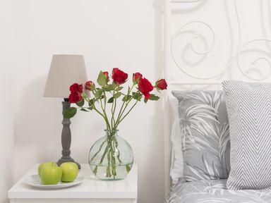 A bedside table with a lamp a vase of red roses and a plate of green apples next to a bed with patterned pillows at Wheal Jewel Lodge in Wendron near Helston