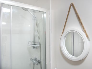 A bathroom shower area with a handheld showerhead enclosed by a glass door and a round mirror hanging on the wall by a rope at Wheal Jewel Lodge in Wendron near Helston