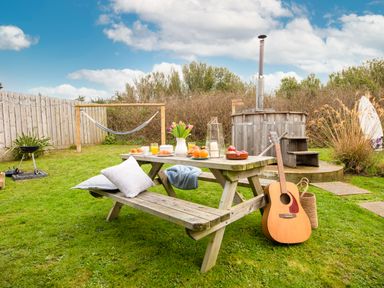An outdoor garden area with a wooden picnic table set with food and drinks a guitar resting against the table a hammock and a wooden hot tub in the background at Wheal Jewel Lodge in Wendron near Helston