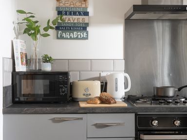 A kitchen countertop with a microwave, toaster, kettle, bread on a cutting board, a pot on the stove, and decorative plants at Wheal Prosper Lodge in Wendron near Helston
