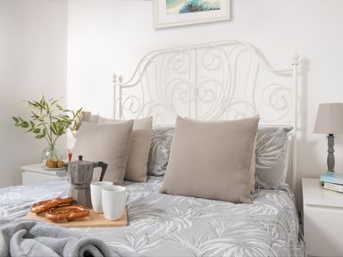 A bed with beige pillows grey and white leaf patterned bedding a tray with a coffee pot two white mugs and pretzels a plant on a bedside table and a lamp at Wheal Prosper Lodge Wendron near Helston