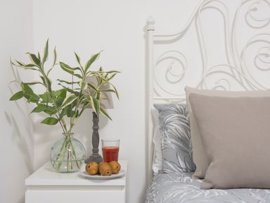 A bedside table with a plant in a glass vase a lamp a plate with fruit and a glass of juice next to a bed with patterned bedding and pillows at Wheal Prosper Lodge Wendron near Helston