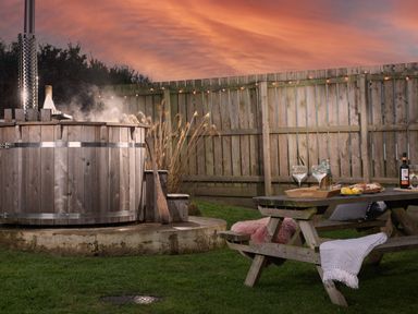 An outdoor wooden hot tub with steam next to a wooden picnic table with drinks and snacks in a fenced garden at Wheal Prosper Lodge in Wendron near Helston