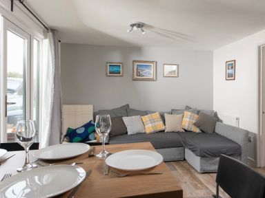 A dining table set with plates and glasses next to a grey sectional sofa with cushions and framed pictures on the wall at Wheal Prosper Lodge in Wendron near Helston