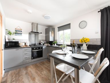 A kitchen with grey cabinets a stove a microwave a table set for three and a window with blinds at Wheal Rose Lodge Wendron near Helston