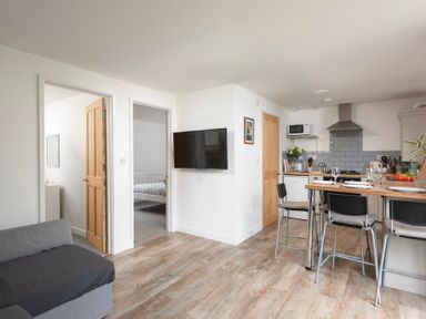 An open plan kitchen and living area with a dining counter set with plates and glasses a wall-mounted television and a view into a bedroom at Wheal Rose Lodge in Wendron near Helston