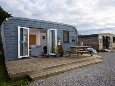 A wooden deck with picnic table and plants outside a small blue cabin with open doors at Wheal Tor Lodge Wendron near Helston