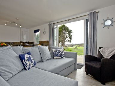 A living area with a light gray sectional sofa with decorative pillows and a dark armchair next to large sliding glass doors overlooking a garden at Wheal Tor Lodge Wendron near Helston