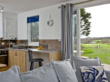 A kitchen corner with a wooden cabinet and countertop with a black chair and a plate of fruit next to a window and glass door with curtains and a gray sofa with cushions in the foreground at Wheal Tor Lodge in Wendron near Helston