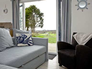 A living room with a gray sofa and crab cushion next to a dark armchair with a pink cloth beside a sliding glass door showing an outdoor view at Wheal Tor Lodge in Wendron near Helston
