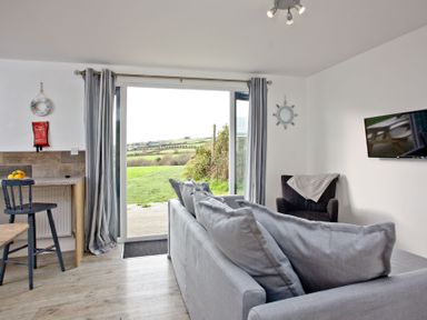 A living area with a gray sofa and pillows a black armchair with a blanket wall-mounted tv a wooden counter with chairs and sliding glass doors showing outdoor greenery at Wheal Tor Lodge in Wendron near Helston