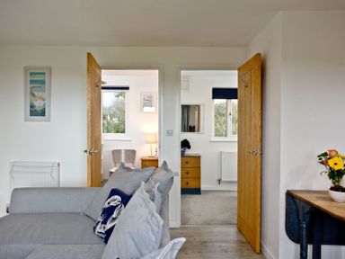 A room with a gray sofa facing two open wooden doors showing a chair and a dresser with windows behind at Wheal Tor Lodge in Wendron near Helston
