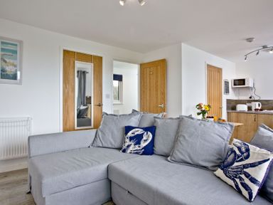 A living room with a gray sectional sofa and decorative pillows near wooden doors and a kitchen area at Wheal Tor Lodge in Wendron near Helston