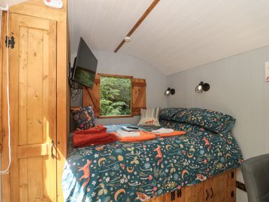 A bedroom with a bed covered in patterned bedding a wooden wardrobe and a wall-mounted TV at The Foxes Den Shepherds Hut Oldbury-On-Severn
