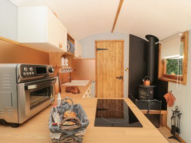 A kitchen with a countertop oven, an electric stove, a basket with muffins, white cabinets, a wooden door, a wood-burning stove, and a window at The Foxes Den Shepherds Hut in Oldbury-On-Severn