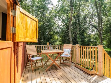 A wooden deck with two chairs a table with wine glasses and a bottle surrounded by trees at The Foxes Den Shepherds Hut in Oldbury-On-Severn