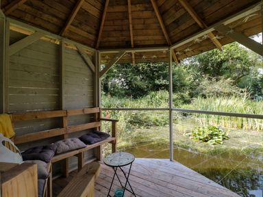 A wooden gazebo with bench seating and cushions overlooking a pond with plants at The Foxes Den Shepherds Hut in Oldbury-On-Severn
