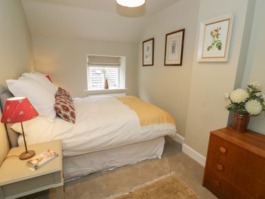 A bedroom with a bed pillows and a yellow blanket a bedside table with a lamp and book a window with a vase of flowers and a wooden chest of drawers with a flower pot at The Bindery in Broadway