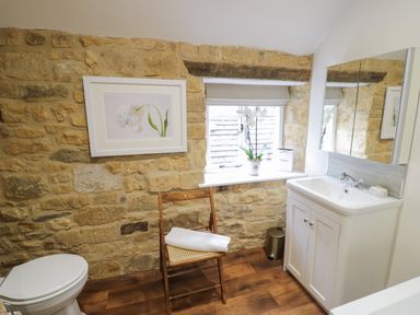A bathroom with a toilet sink wooden chair and a window on a stone wall at The Bindery in Broadway