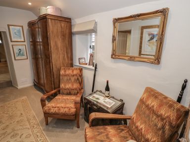 A seating area with two patterned armchairs a wooden wardrobe a decorative mirror and a table with a book and wine bottle at The Bindery in Broadway