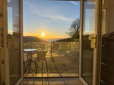 An open glass door leading to a patio with a table and chair overlooking hills and a sunset at Crogal Farmhouse in New Quay