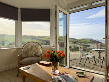A sitting area with a cushioned chair a wooden table with flowers magazines and a game set glass doors opening to an outdoor patio with chairs and a table overlooking green hills at Crogal Farmhouse in New Quay