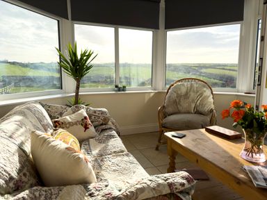 A living room with a sofa with cushions a wooden chair a wooden coffee table with a vase of orange flowers and a plant near large windows at Crogal Farmhouse in New Quay