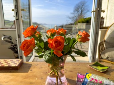 A vase with orange roses on a wooden table in a room with open doors leading to an outdoor patio at Crogal Farmhouse in New Quay