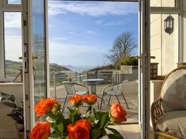 An open doorway leading to a patio with a table and two chairs and a view of hills and water with orange flowers in the foreground at Crogal Farmhouse in New Quay