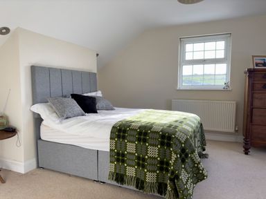 A bedroom with a grey upholstered bed with pillows and a green patterned blanket near a window and a wooden dresser at Crogal Farmhouse in New Quay