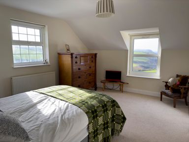 A bedroom with a bed covered by a green patterned blanket a wooden chest of drawers a small table with a TV and a chair with a teddy bear at Crogal Farmhouse in New Quay