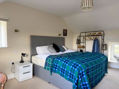 A bedroom with a double bed covered by a blue and green patterned blanket a bedside table with a lamp and a clothes rack with hangers at Crogal Farmhouse in New Quay