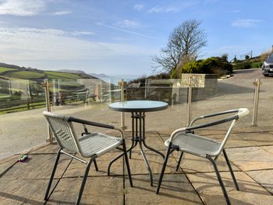 An outdoor patio with two woven chairs and a round glass table overlooking hills and a sea view at Crogal Farmhouse in New Quay