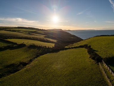 A coastal landscape with green fields hills and the sun near the horizon at Crogal Farmhouse in New Quay