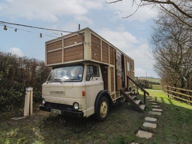 A converted vintage truck with wooden paneling and stairs leading to a door parked on grass with trees and string lights at Sea the Stars in New Quay