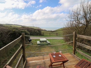 A wooden deck with two chairs and a table with two glasses overlooking a picnic table and green fields with a view of the sea at Sea the Stars in New Quay