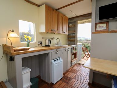 A small kitchen with wooden cabinets a sink a kettle a lamp and flowers on the counter with an open door leading to an outdoor seating area at Sea the Stars in New Quay