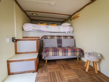 A small room with a bed on a raised platform a couch with a plaid blanket and gray cushions wooden flooring stairs a small round stool with magazines