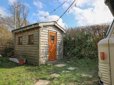 A small wooden shed with a brown door and two windows in a grassy garden with string lights overhead and a white vehicle partially visible at Sea the Stars in New Quay
